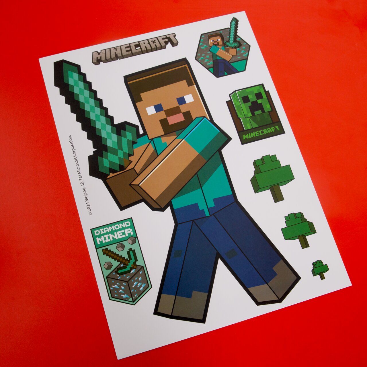 Schräg von oben fotografierter Minecraft-Stickerbogen auf rotem Hintergrund mit Steve und Diamantschwert sowie Creeper- und Baum-Motiven