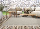 Outdoor-Sitzgruppe mit beigem Teppich, Holztischen und beigen Sofas vor einer Steinmauer, von vorne betrachtet.