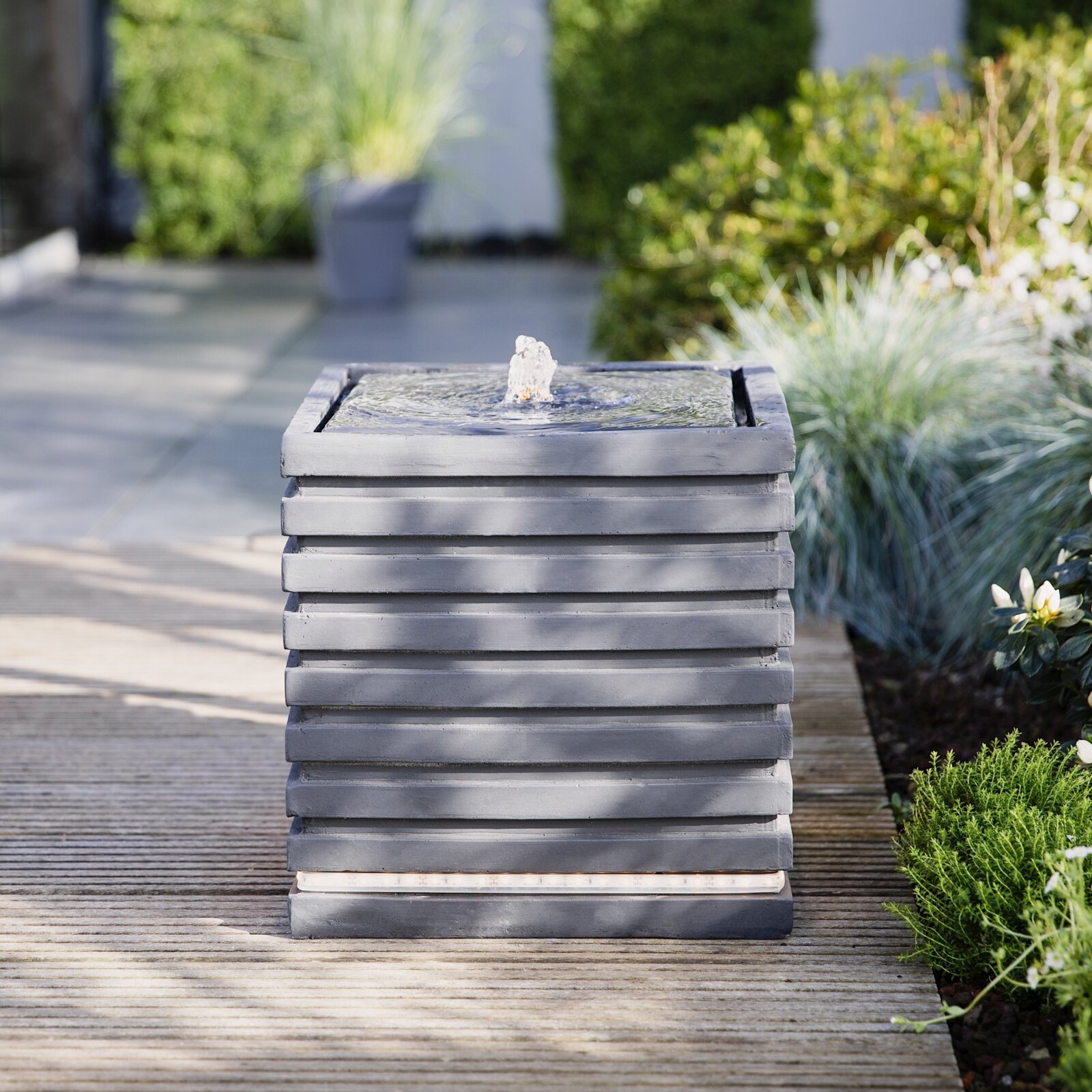 Moderner, quadratischer Garten-Standbrunnen aus grauem Stein/Beton mit kleiner Wasserfontäne oben, frontale Perspektive auf einer Holzterrasse im grünen Garten.