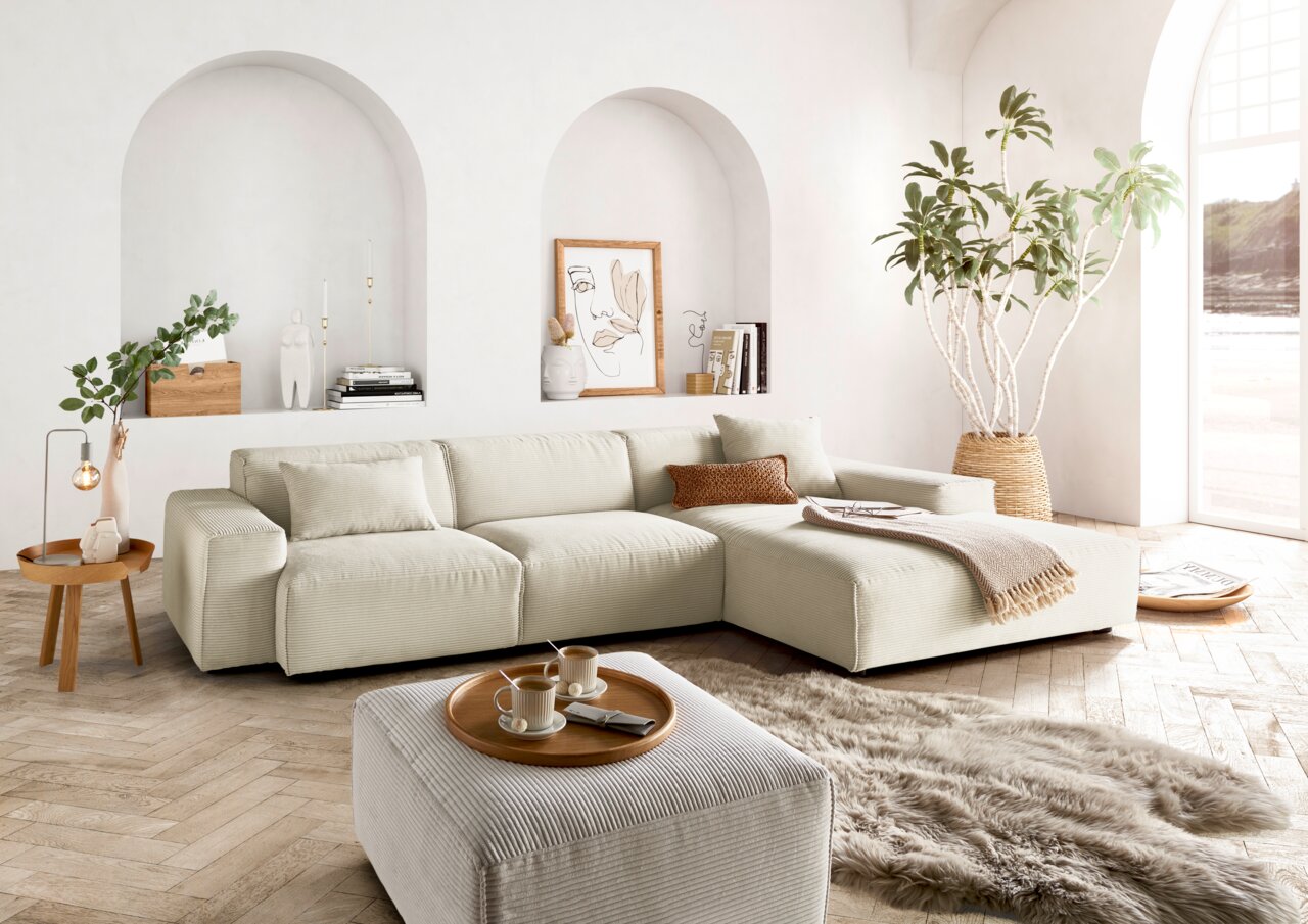 Lounge Handmade Hoekbank LH-EDINA Helle, moderne Wohnzimmer-Ecke mit einer großen, beigen Rundecke und passendem Hocker, umgeben von stilvoller Dekoration und Pflanzen. Perspektive von vorne.