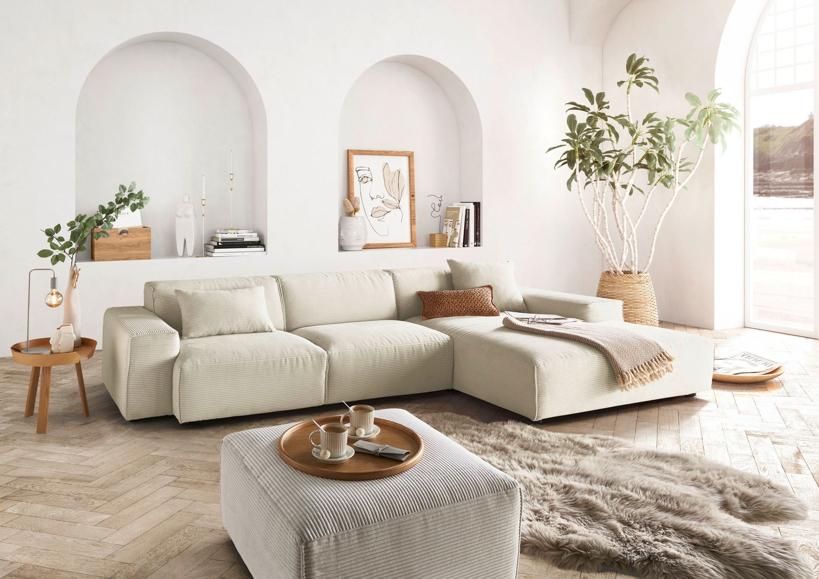 Helle, moderne Wohnzimmer-Ecke mit einer großen, beigen Rundecke und passendem Hocker, umgeben von stilvoller Dekoration und Pflanzen. Perspektive von vorne.