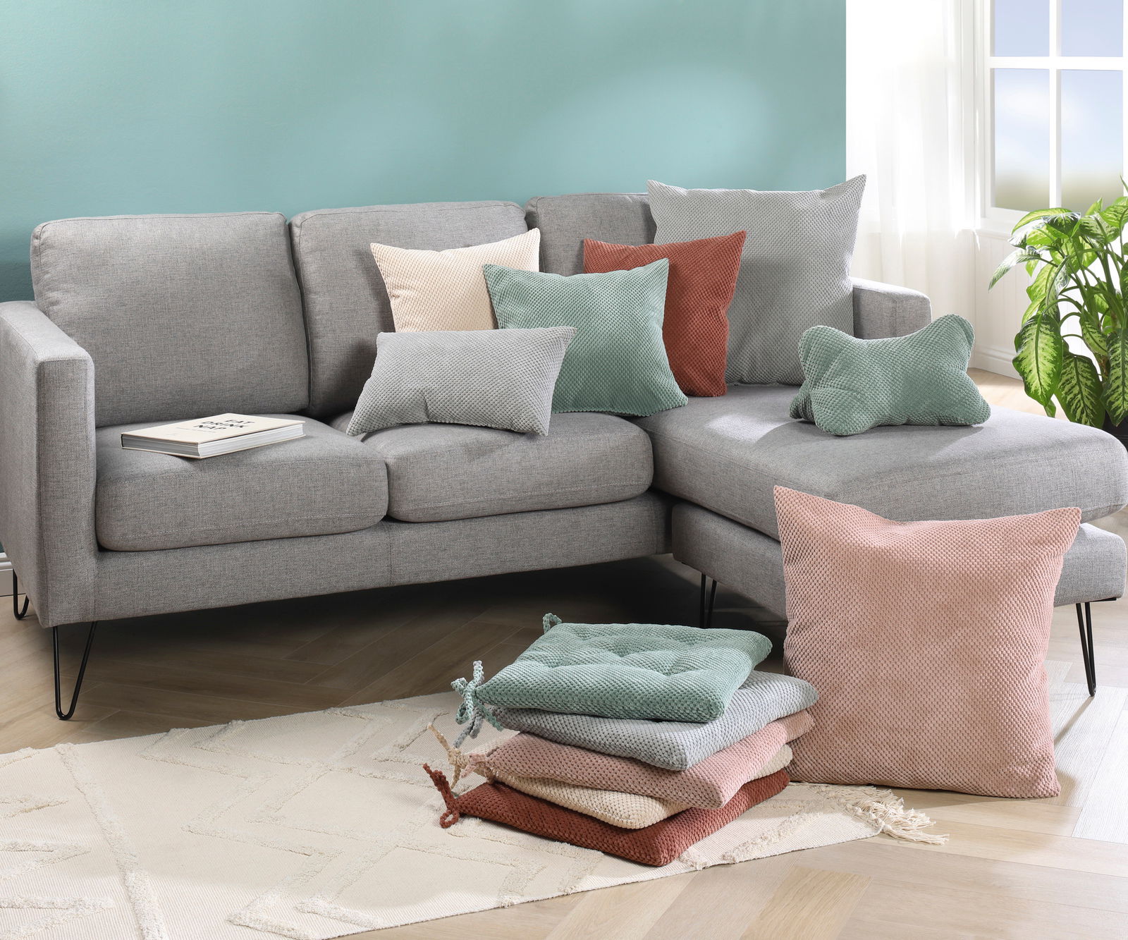 Ein modernes Wohnzimmer mit einem grauen Sofa, dekoriert mit verschiedenen Kissen in Pastellfarben. Die Kissenhüllen sind in den Farben Rosa, Grün und Beige gehalten. Im Vordergrund ist ein Stapel von Kissen auf einem hellen Teppich zu sehen. Die Perspekt