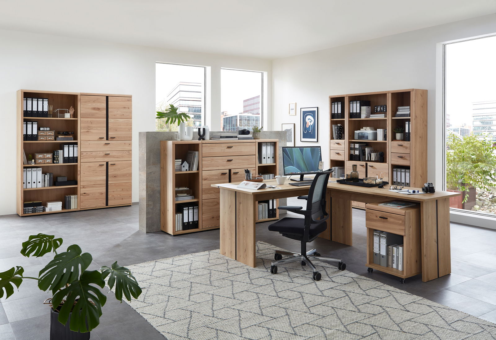 Moderne Büromöbel in einem hellen Raum mit großen Fenstern, bestehend aus Schreibtischen, Aktenschränken und Regalen aus Holz. Perspektive von vorne links.