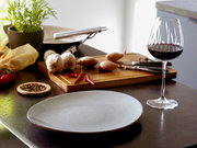 Hellgrauer, gesprenkelter Speiseteller auf dunkler Küchenarbeitsplatte, im Vordergrund aus leicht schräger Frontansicht; im Hintergrund Schneidebrett mit Knoblauch, Schalotten und Chili sowie ein Glas Rotwein.
