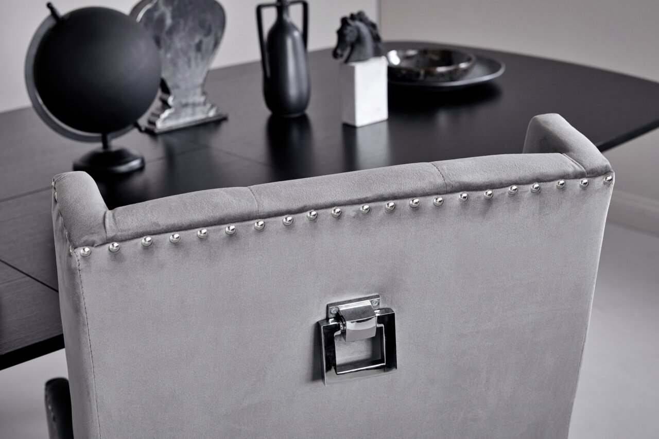 Achterkant van een elegante grijze stoel met zilveren studs en een decoratieve handgreep, voor een zwarte tafel met decoratieve voorwerpen.