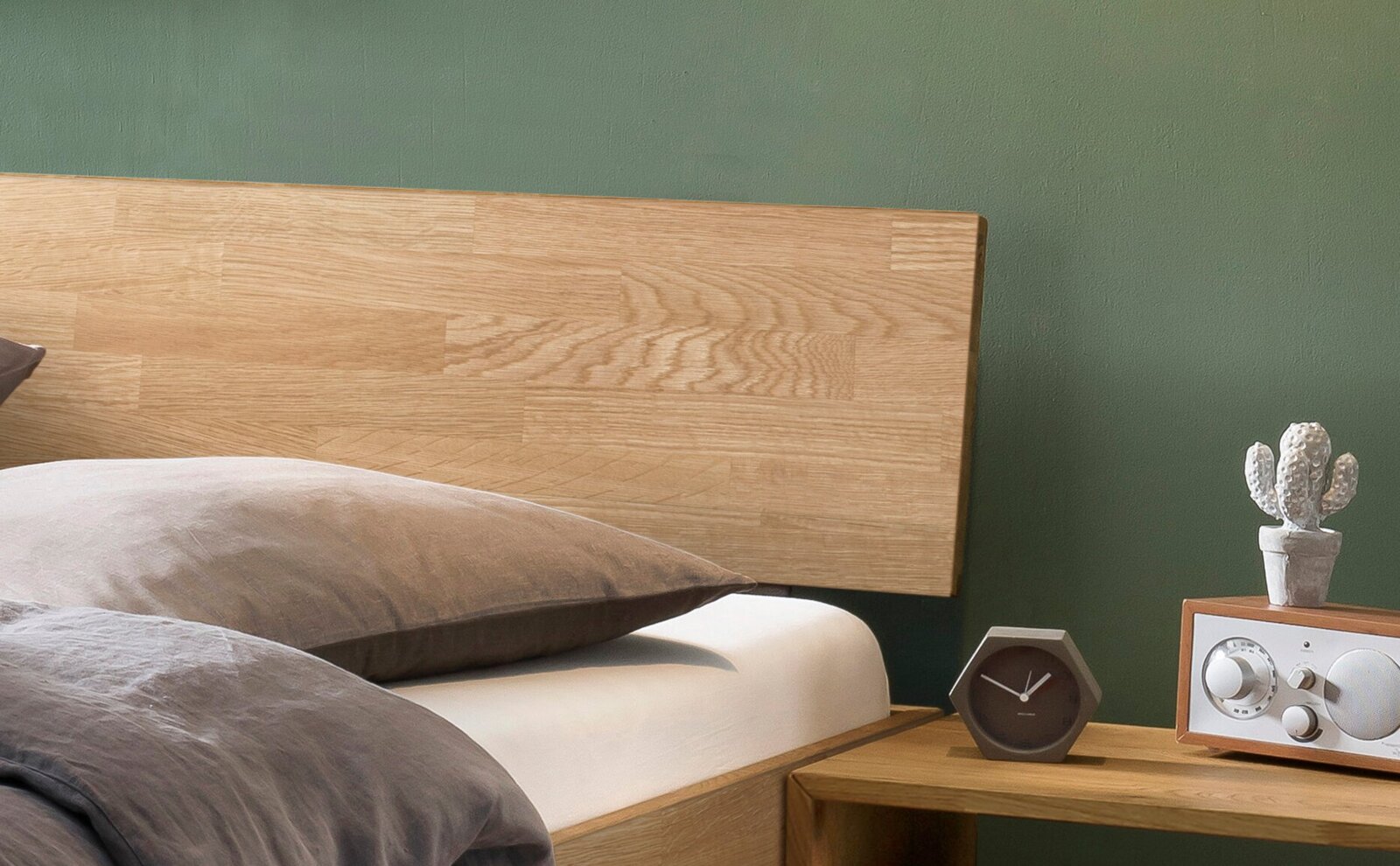 Nahaufnahme eines Bettes mit hellem Holz-Kopfteil und grauer Bettwäsche neben einem Nachttisch mit Uhr, Radio und kleiner Kaktus-Deko vor grüner Wand, seitliche Perspektive