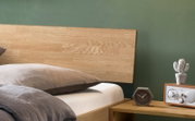 Nahaufnahme eines Bettes mit hellem Holz-Kopfteil und grauer Bettwäsche neben einem Nachttisch mit Uhr, Radio und kleiner Kaktus-Deko vor grüner Wand, seitliche Perspektive