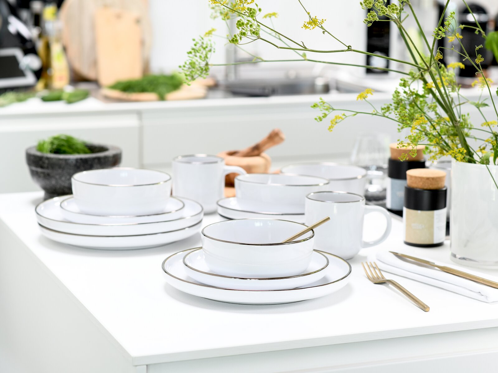 Modernes weißes Geschirrset mit feinem Goldrand (Teller, Schalen und Becher) auf weißer Kücheninsel, mit goldenem Besteck und Blumen im Hintergrund, aufgenommen in leichter Seitenansicht von vorne