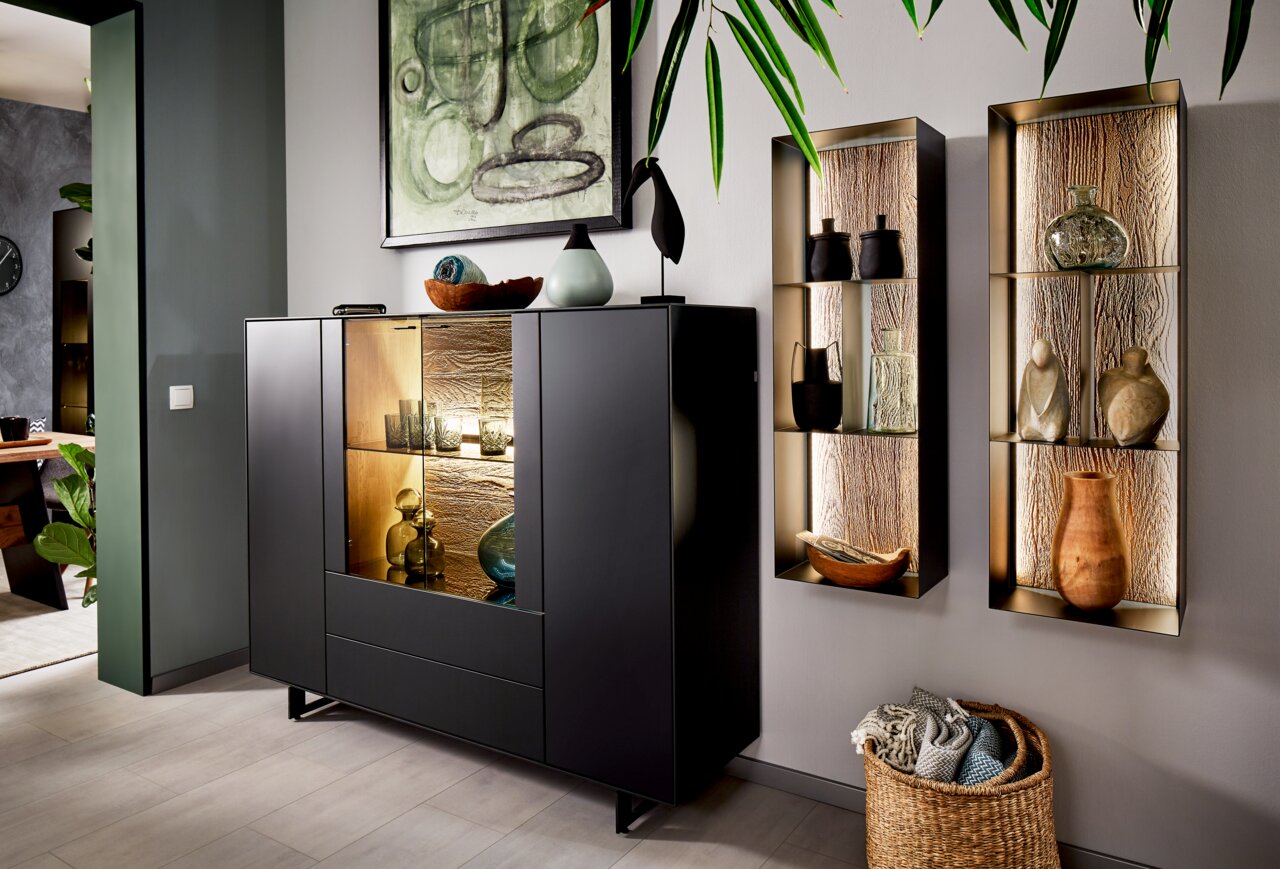 Valnatura Hoog dressoir LIVINO Modernes Highboard in dunklem Design mit Glasvitrine, dekoriert mit Keramik und Glasobjekten, aus seitlicher Perspektive fotografiert.