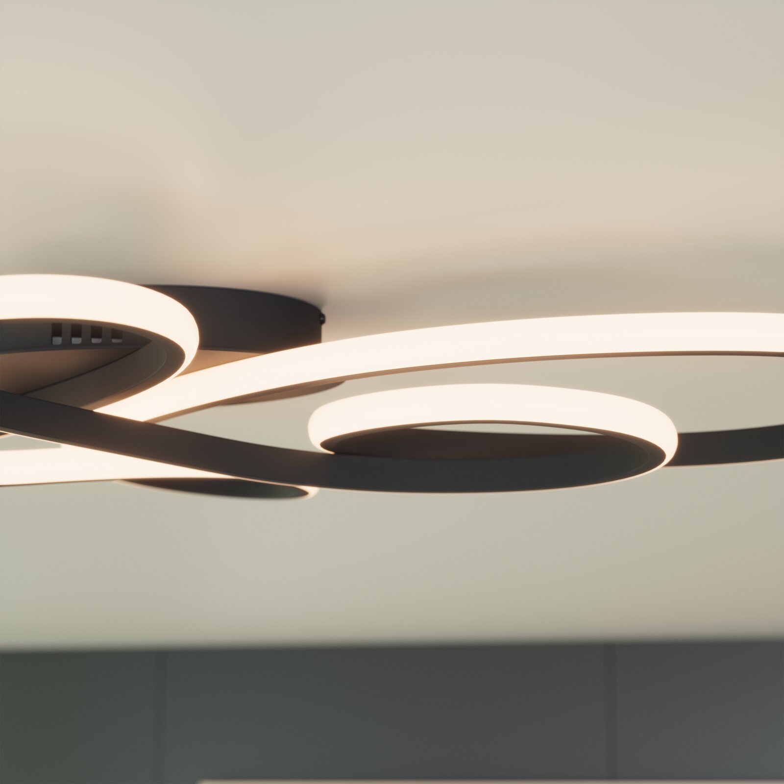 Moderne LED-Deckenleuchte in Schwarz mit geschwungenen, ringförmigen Lichtbändern an heller Decke, warmweiß leuchtend, fotografiert aus Untersicht in leichter Schrägperspektive.