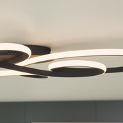Moderne LED-Deckenleuchte in Schwarz mit geschwungenen, ringförmigen Lichtbändern an heller Decke, warmweiß leuchtend, fotografiert aus Untersicht in leichter Schrägperspektive.