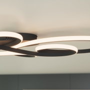 Moderne LED-Deckenleuchte in Schwarz mit geschwungenen, ringförmigen Lichtbändern an heller Decke, warmweiß leuchtend, fotografiert aus Untersicht in leichter Schrägperspektive.