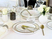 Elegant gedeckter Tisch mit weißen Tellern mit goldfarbenem Rand, goldfarbenem Besteck, Wein- und Wassergläsern sowie weißen Rosen in Vasen, perspektivisch aus leicht erhöhter schräger Vorderansicht.