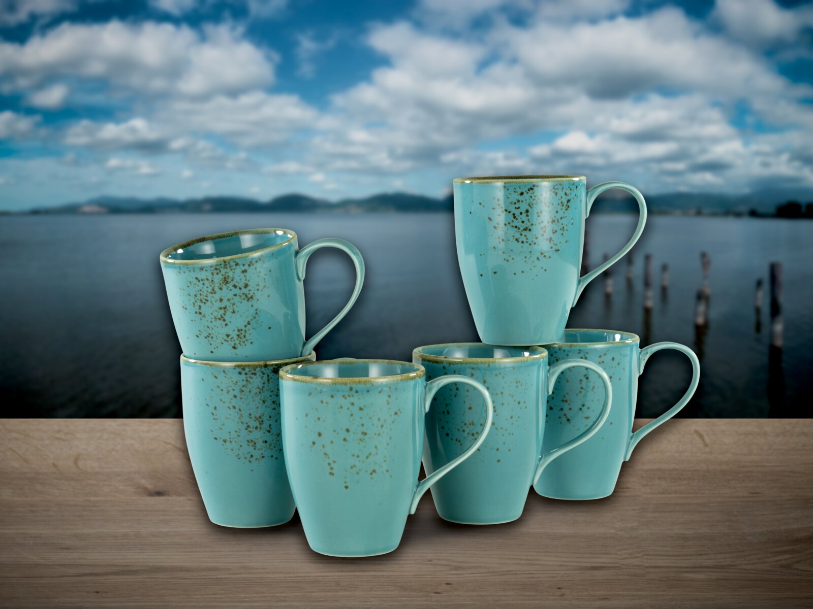 Fünf türkisfarbene, gesprenkelte Keramik-Kaffeebecher mit Henkel auf Holztisch vor See- und Himmel-Hintergrund, fotografiert aus frontaler Perspektive mit leichter Aufsicht.