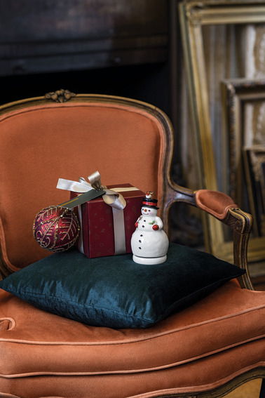 Drehende Spieluhr in Form eines Schneemanns, auf einem grünen Kissen auf einem orangefarbenen Sessel platziert, mit einem Geschenk und einer Weihnachtskugel daneben, seitliche Perspektive.