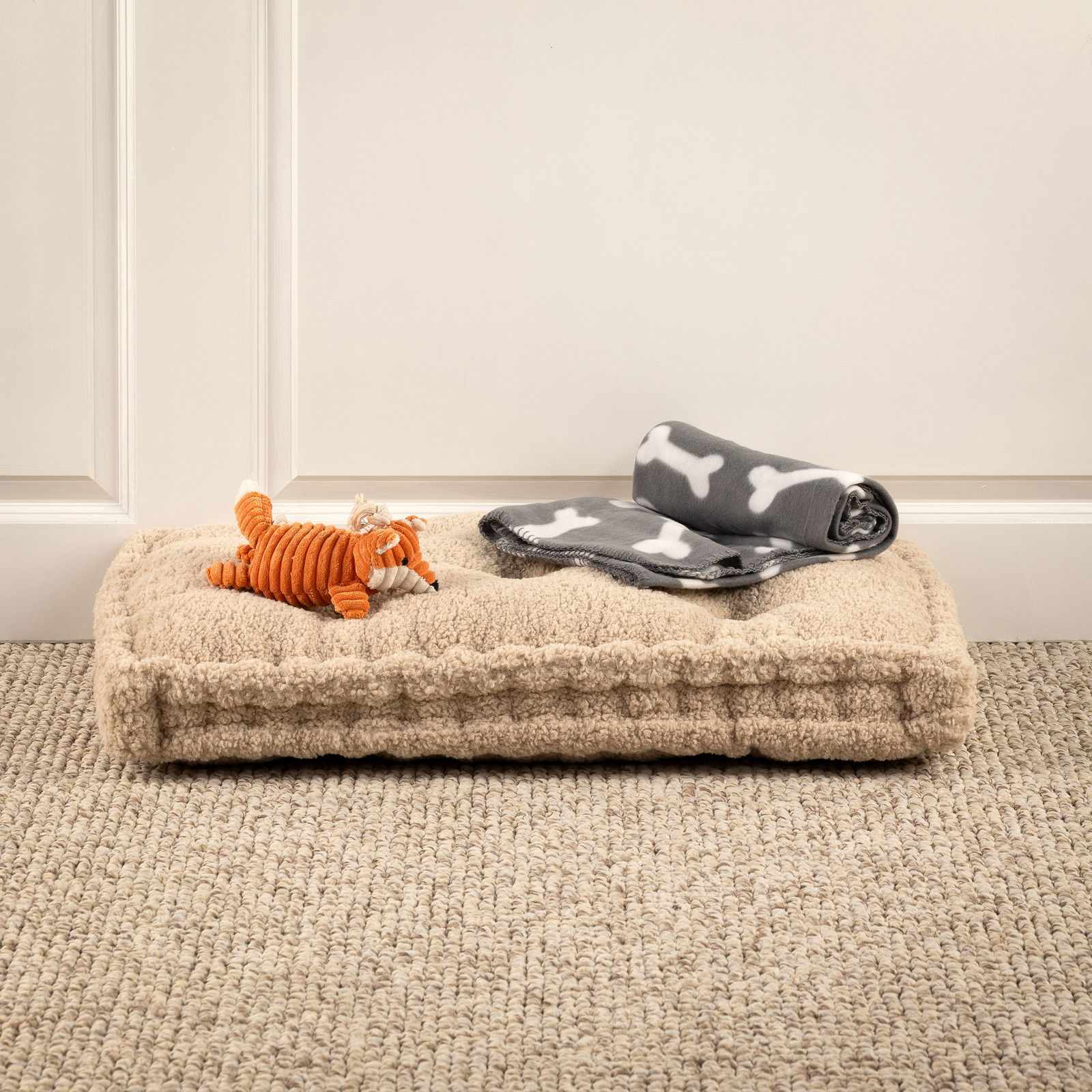 Hundespielzeug-Set mit orangefarbenem Plüschfuchs und grauer Decke mit Knochenmuster auf beigem Hundebett, seitliche Perspektive