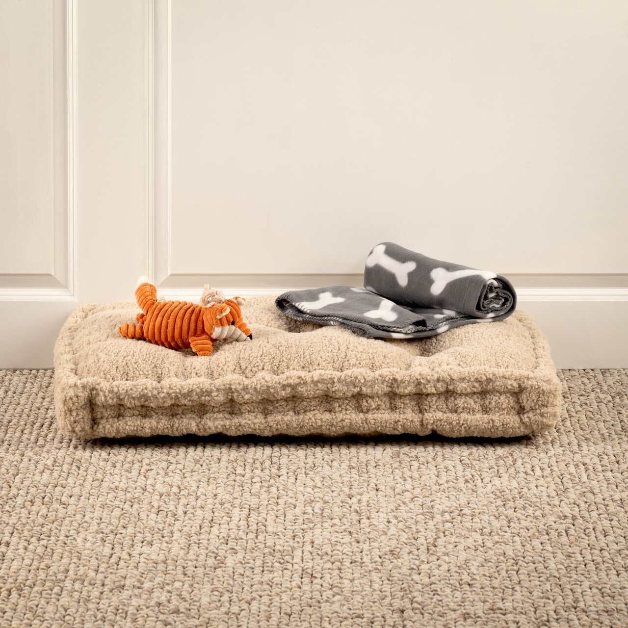 Hundespielzeug-Set mit orangefarbenem Plüschfuchs und grauer Decke mit Knochenmuster auf beigem Hundebett, seitliche Perspektive