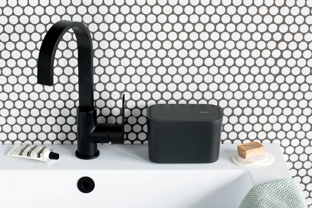 Brabantia Abfallbehälter MINDSET Moderner schwarzer Abfallbehälter auf einem weißen Waschbecken, neben einem schwarzen Wasserhahn, mit einer stilvollen, gepunkteten Wand im Hintergrund. Perspektive von vorne.