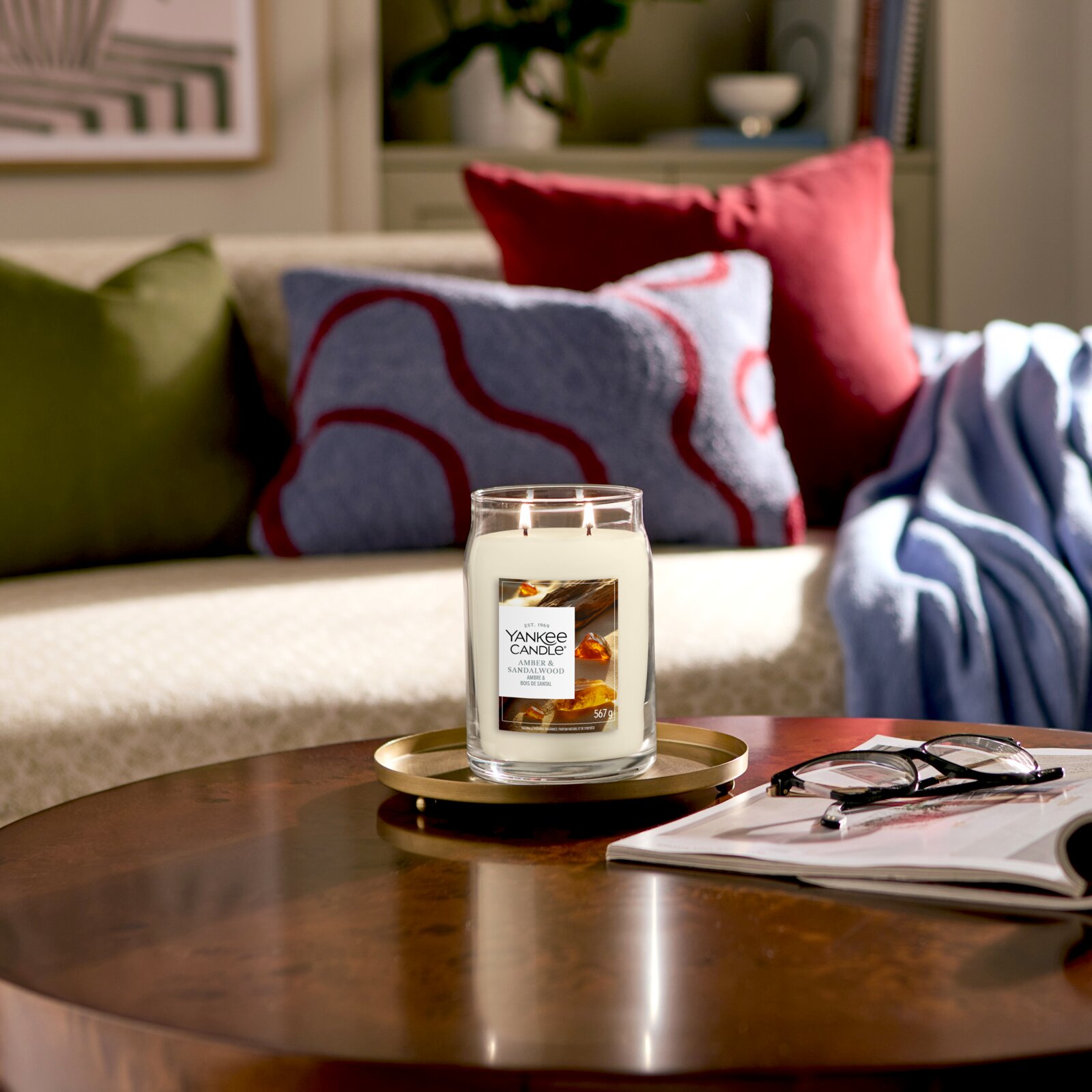 Frontalansicht einer brennenden Yankee Candle Amber & Sandalwood mit zwei Dochten im großen Glas, dekorativ auf einem goldfarbenen Tablett im gemütlichen Wohnzimmer platziert