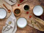 Drei Keramikschalen mit weißer Innenfläche und grünem Rand auf rustikalem Holztisch, umgeben von Holzbrettchen, Löffeln, Oliven und Cherrytomaten, fotografiert in Draufsicht (Flatlay).