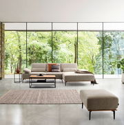 Großer Valnatura Hocker SPRINT in einem modernen Wohnzimmer mit großen Fenstern und Blick auf einen grünen Wald, Perspektive von vorne.