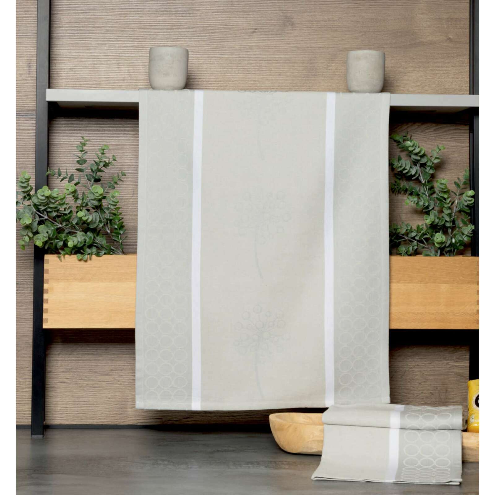 Beige Geschirrtuch mit dezentem Muster, aufgehängt vor einer Holzwand mit Pflanzen im Hintergrund, Vorderansicht