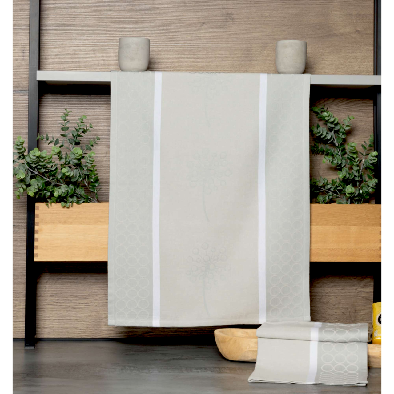 Beige Geschirrtuch mit dezentem Muster, aufgehängt vor einer Holzwand mit Pflanzen im Hintergrund, Vorderansicht