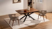 Moderne houten eettafel met donkerbruin blad en zwart X-vormig onderstel, gefotografeerd in schuin vooraanzicht in een lichte eetkamer met twee gestoffeerde stoelen op een beige vloerkleed.