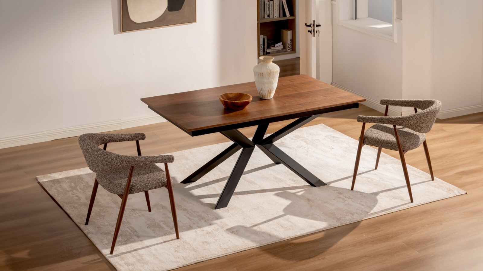 Moderne houten eettafel met donkerbruin blad en zwart X-vormig onderstel, gefotografeerd in schuin vooraanzicht in een lichte eetkamer met twee gestoffeerde stoelen op een beige vloerkleed.