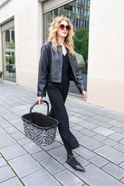 Frau in schwarzer Kleidung trägt einen Einkaufskorb mit Leopardenmuster auf der Straße, seitliche Perspektive.