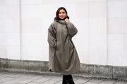 Frontansicht: Frau trägt einen oversized, knielangen Regenmantel/Parka in Taupe mit Kapuze und Seitentaschen, Outdoor vor heller Wand.