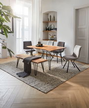 Moderner Esstisch mit Holzplatte und schwarzen Metallbeinen, umgeben von grauen Stühlen und einer Bank, in einem hellen Raum mit Teppich und Dekorationen, seitliche Perspektive.