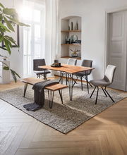 Moderner Esstisch mit Holzplatte und schwarzen Metallbeinen, umgeben von grauen Stühlen und einer Bank, in einem hellen Raum mit Teppich und Dekorationen, seitliche Perspektive.