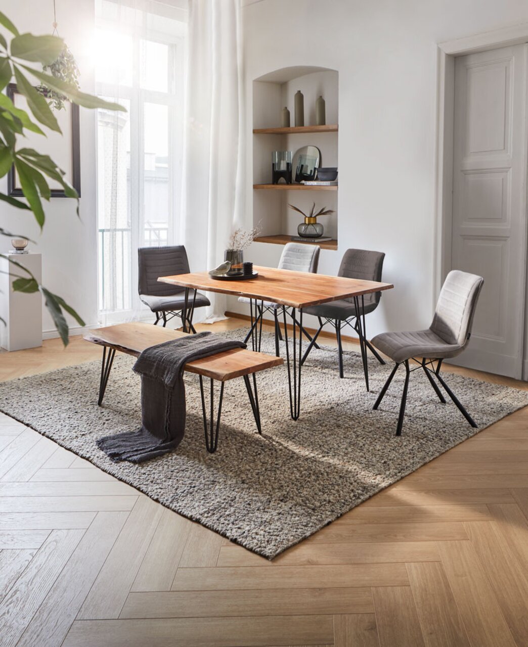 Moderner Esstisch mit Holzplatte und schwarzen Metallbeinen, umgeben von grauen Stühlen und einer Bank, in einem hellen Raum mit Teppich und Dekorationen, seitliche Perspektive.