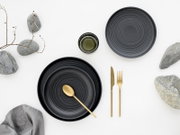 Dunkles, gerilltes Keramikgeschirr-Set mit zwei Tellern und kleiner Schale, dazu goldfarbenes Besteck auf weißem Hintergrund mit Deko-Steinen, Drahtzweig und Stoffserviette, aus der Draufsicht (Top-Down).