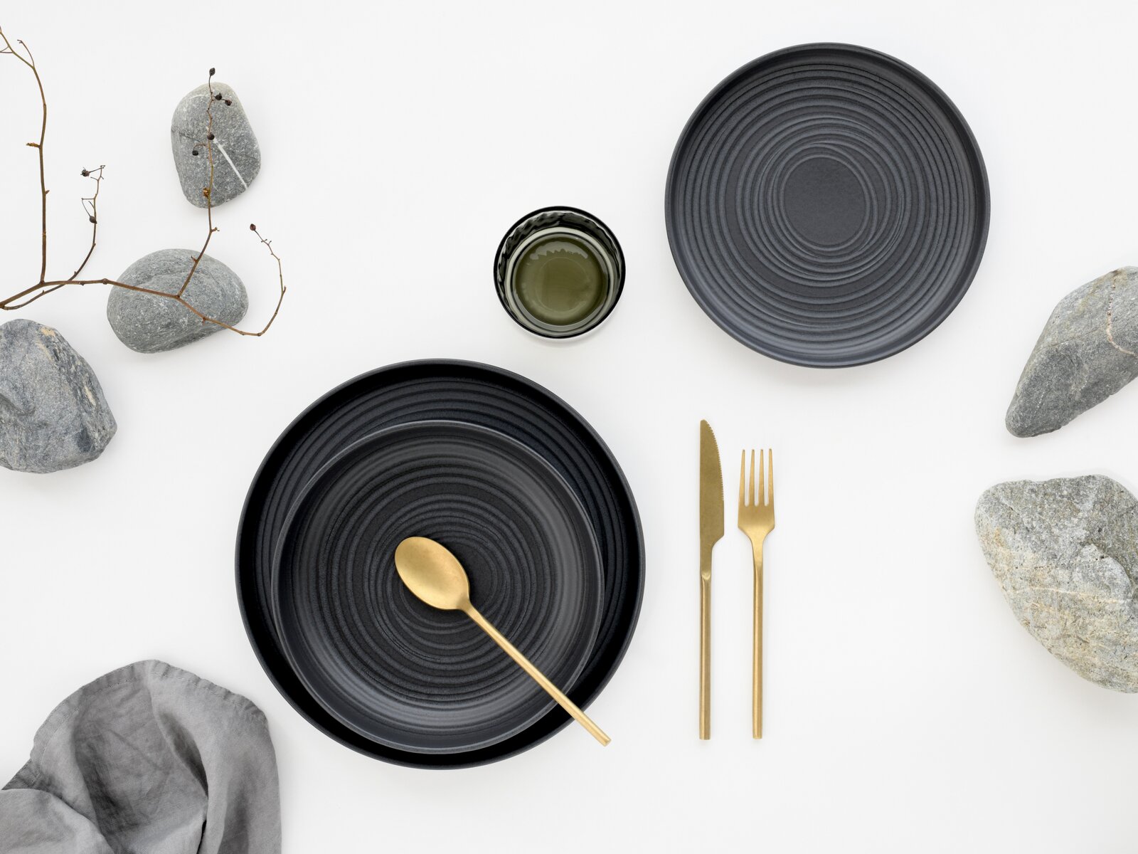 Dunkles, gerilltes Keramikgeschirr-Set mit zwei Tellern und kleiner Schale, dazu goldfarbenes Besteck auf weißem Hintergrund mit Deko-Steinen, Drahtzweig und Stoffserviette, aus der Draufsicht (Top-Down).