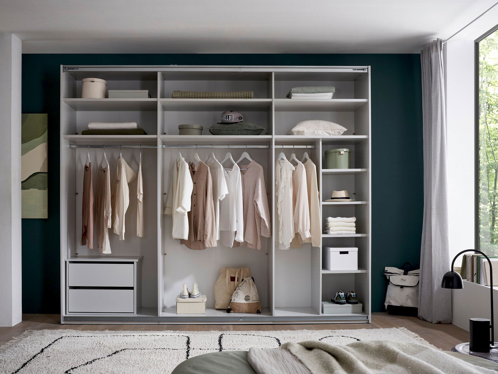 Offener, weißer Kleiderschrank mit Regalböden, Kleiderstangen, Schubladen und ordentlich aufgehängter Kleidung in einem Schlafzimmer, frontale Perspektive.