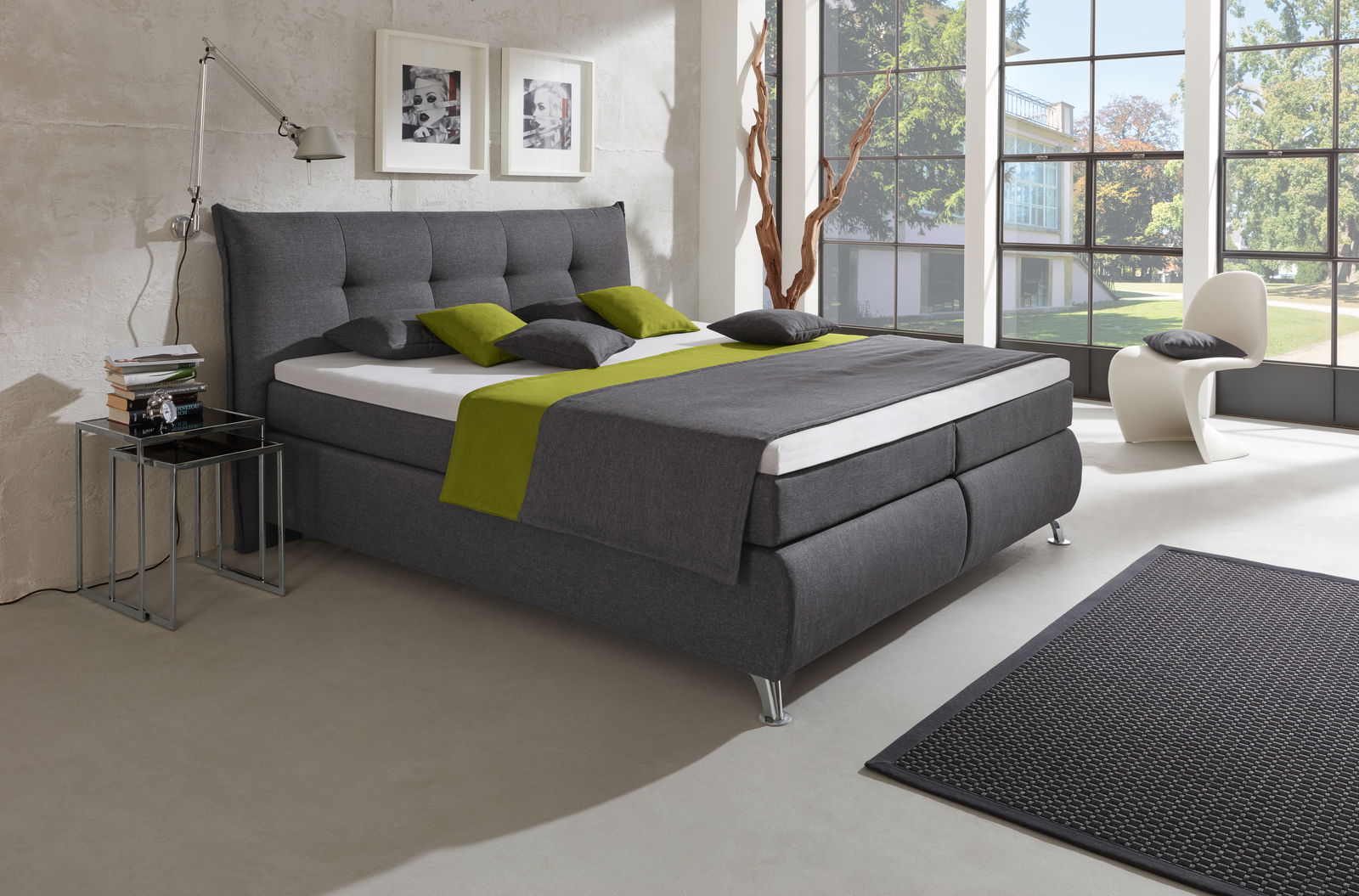 Modernes Boxspringbett in einem hellen Raum mit großen Fenstern, grauem Stoffbezug und grünen Akzenten, seitliche Perspektive.