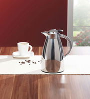 Edelstahl-Isolierkanne Signo auf einem Tisch, seitliche Perspektive, neben einer Tasse Kaffee.