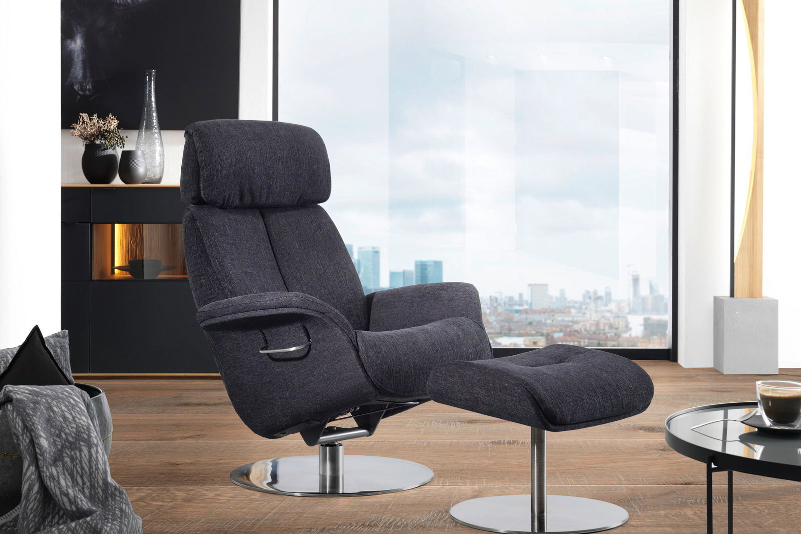 Welnova Relaxfauteuil 7602 BALANCE Moderner Relaxsessel mit Hocker in dunklem Stoffbezug, seitliche Perspektive, vor einem großen Fenster mit Stadtblick.