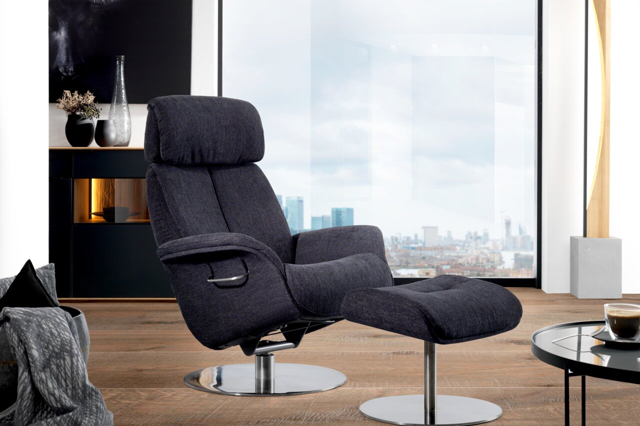 Welnova Relaxsessel 7602 BALANCE Moderner Relaxsessel mit Hocker in dunklem Stoffbezug, seitliche Perspektive, vor einem großen Fenster mit Stadtblick.