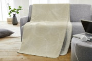 Beige jacquarddeken met cirkelvormig patroon, gedrapeerd over een grijze bank in een moderne woonkamer, gezien vanuit de zijkant.