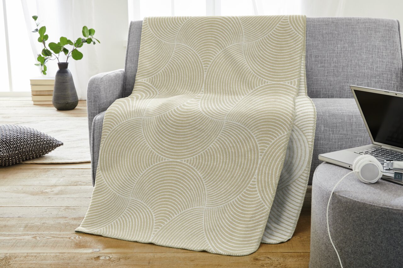 Beige jacquarddeken met cirkelvormig patroon, gedrapeerd over een grijze bank in een moderne woonkamer, gezien vanuit de zijkant.