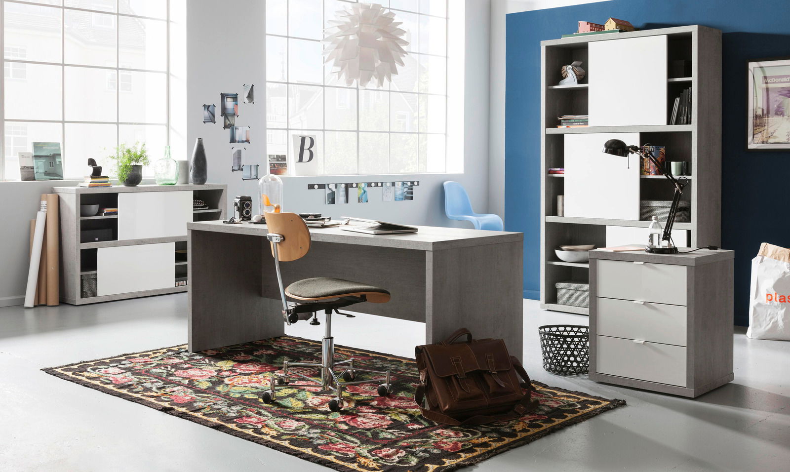 Modernes Büro mit grauem Schreibtisch, Rollcontainer und Hochschrank in einem hellen Raum mit großen Fenstern, seitliche Perspektive.