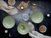 Drei salbeigrüne Keramikschalen mit braunem Rand auf dunkler Tischplatte, dekoriert mit goldfarbenen Löffeln, Holzbrett, Kerzen und Leinentüchern, Draufsicht (Flatlay).