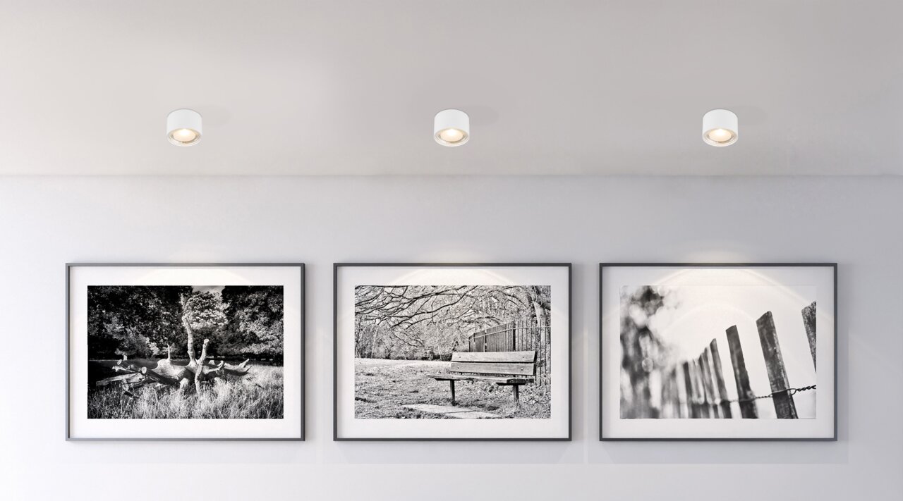 Deckenleuchte aus weißem Metall, von unten betrachtet, beleuchtet eine Wand mit drei schwarz-weißen Fotografien.