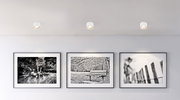 Deckenleuchte aus weißem Metall, von unten betrachtet, beleuchtet eine Wand mit drei schwarz-weißen Fotografien.