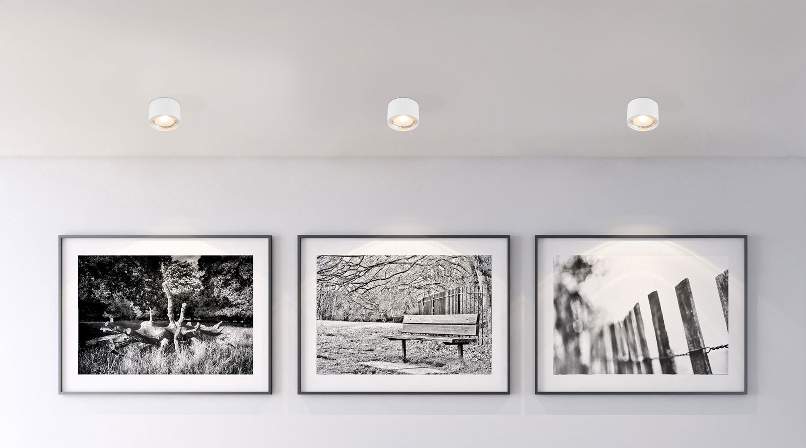 Deckenleuchte aus weißem Metall, von unten betrachtet, beleuchtet eine Wand mit drei schwarz-weißen Fotografien.
