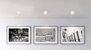 Deckenleuchte aus weißem Metall, von unten betrachtet, beleuchtet eine Wand mit drei schwarz-weißen Fotografien.