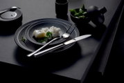 Schwarzes, mattes Teller-Set mit silbernem Messer und Gabel auf dunklem Tisch, mit kleiner Speise angerichtet; Perspektive: leicht schräg von oben (3/4-Ansicht).